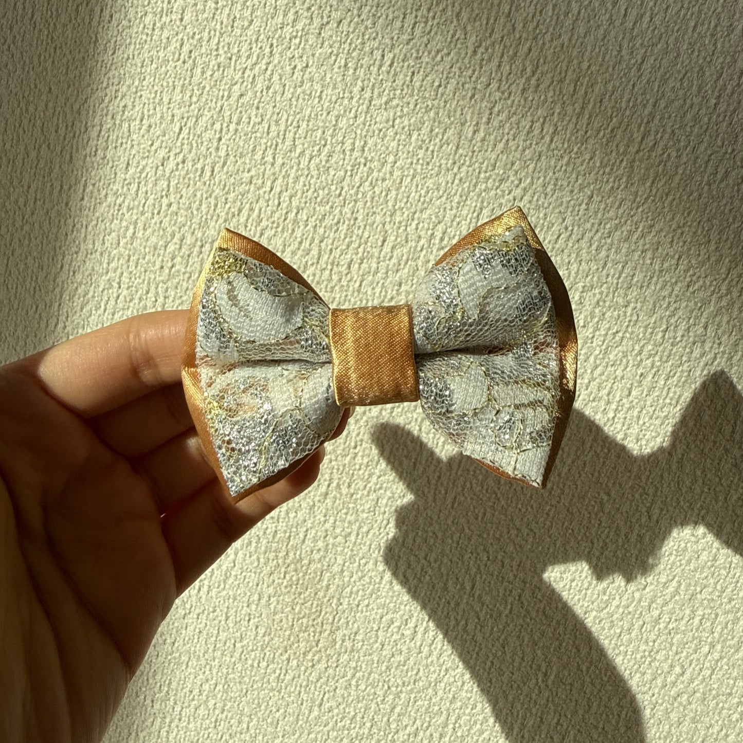 Hand holding a gold satin bow with white floral embroidered layer on top against a textured beige background.