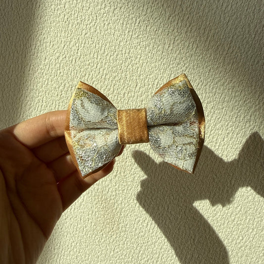 Hand holding a gold satin bow with white floral embroidered layer on top against a textured beige background.
